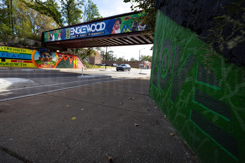 A street-level view beneath a railroad viaduct in Chicago's Englewood neighborhood, featuring a vibrant community mural that reads "Our Englewood: Home, Family, Community." The underpass walls display painted words — "Strength, Resilience, Peace and Growth" — alongside colorful imagery celebrating the neighborhood.