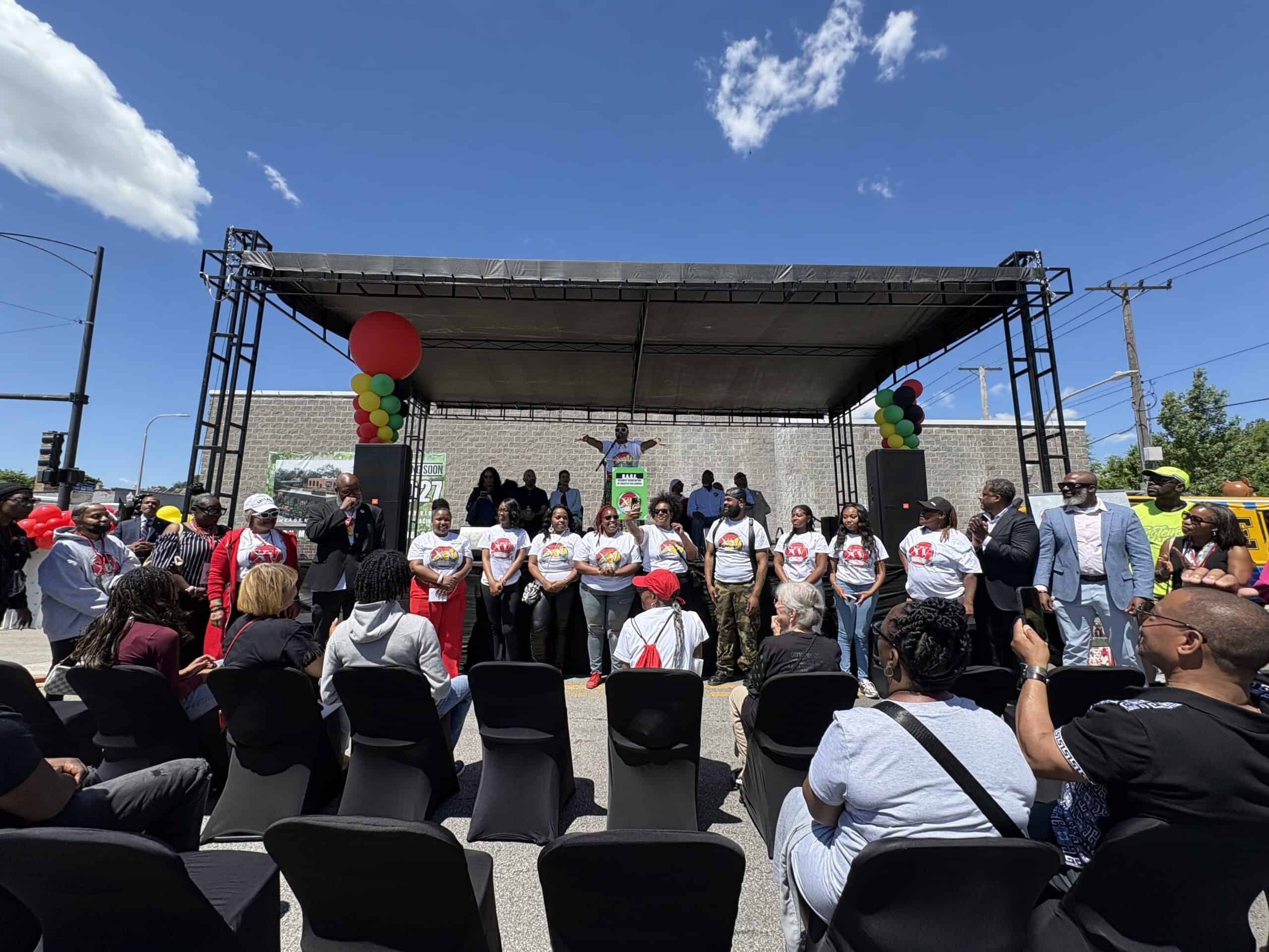 A celebration hosted by R.A.G.E. in Englewood, held on an outdoor stage decorated with red, black, yellow, and green balloon garlands. A group of R.A.G.E. members in matching white T-shirts smile and laugh at the front of the stage while R.A.G.E co-founder and CEO Asiaha Butler gestures at a podium behind them. An audience watches and claps in the foreground under a clear blue sky.