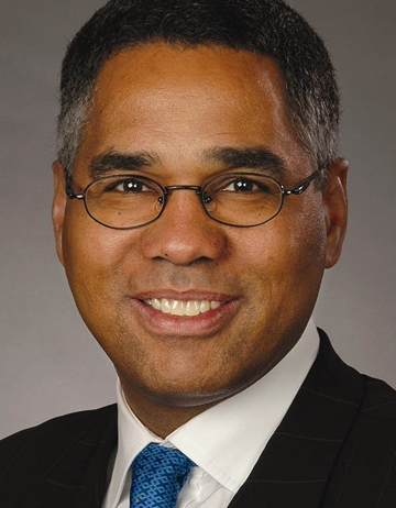 Lyle is smiling against a gray background. He is wearing dark, wire-rimmed glasses, a black jacket over a white-collared shirt and a royal blue patterened tie.