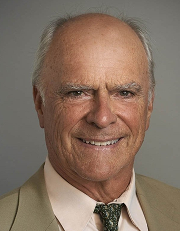 Marshall is picutred against a gray background. He is smiling and wearing a tan jacket over a cream-colored collared shirt with a dark patterned tie.