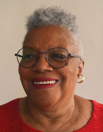 Rita is smiling against a beige background. She is wearing dark-rimmed rounded glasses, gold earrings, and a red sweater.