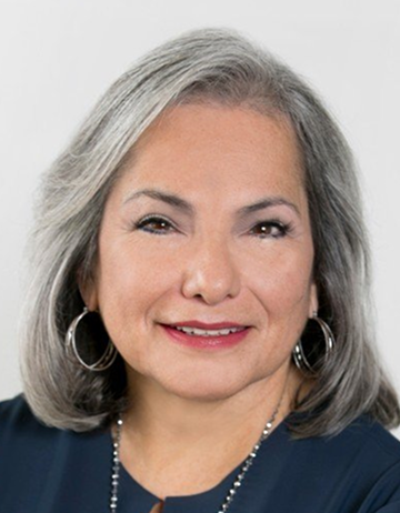 Gloria is smiling against a light backdrop. She is wearing a navy blouse with silver hoop earrings, and a silver necklace