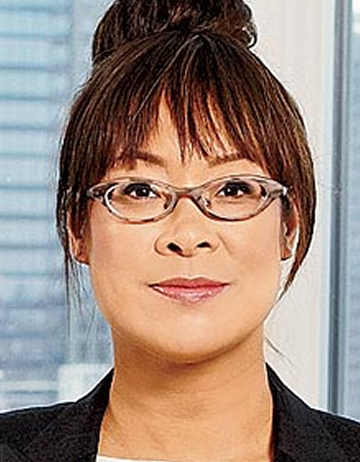 Lisa is pictured against an office window. She has a closed smile and is wearing tortoise shell, cat-style glasses and a black jacket.