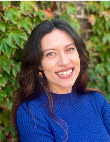 Edith is posing against a background of greenery. She is smiling and wearing a royal blue sweater.