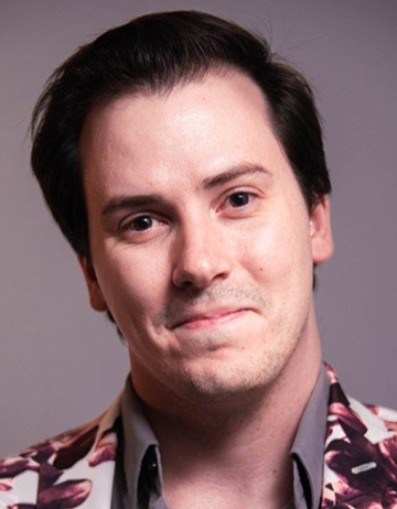 Patrick is smiling against gray background and wearing a white, red, and black patterned shirt