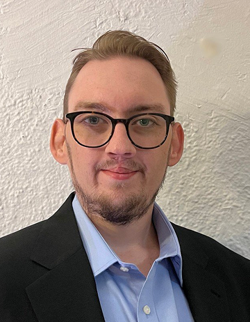 Thomas is posing against a tan backdrop. He is wearing dark rimmed glasses and a dark jacket over a light blue collared shirt.