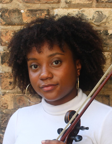 Michelle is posing with a slight smile against a brick wall. She is wearing hoop earrings and is wearing a white turtleneck and holding a violin.