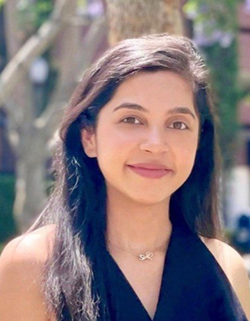 Rupal is posing against a natural background. She has a slight smile and is wearing a sleeveles, v-neck blouse with a necklace.