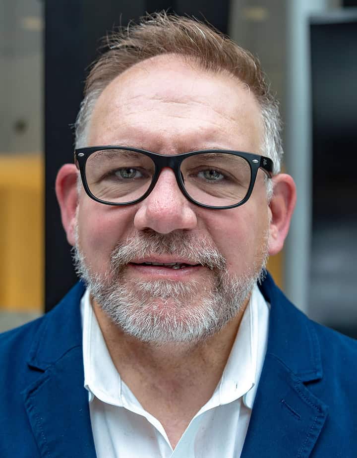 Mark is pictured in an open space with a textured background. He has a slight smile and is wearing black glasses, a navy blue and a white collared shirt.