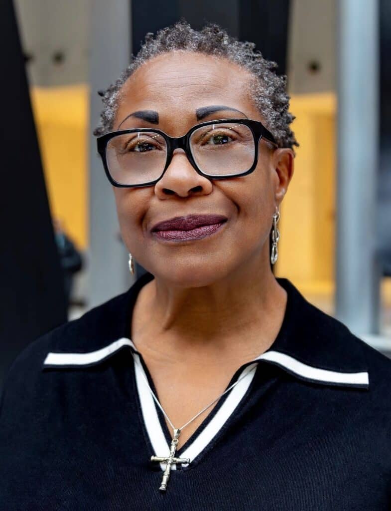 Maudlyne is pictured in an open space with a textured background. She is smiling and wearing black, rimmed glasses and a black sweater with a white stripe on the collar.