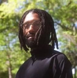 Elijah is pictured in an outdoor setting with trees in the background. He has a serious expression and is wearing a dark shirt.