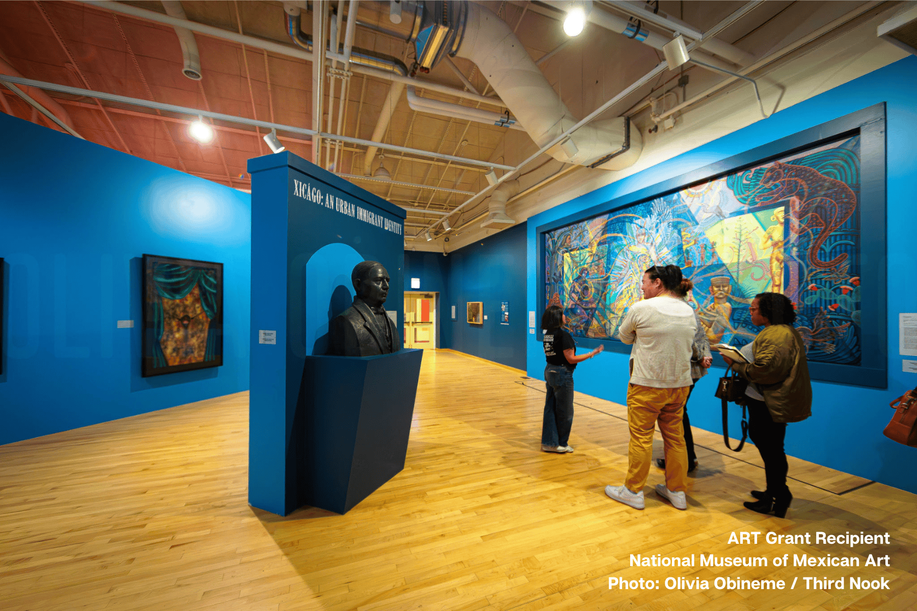Museum visitors viewing exhibition at ART grant recipient National Museum of Mexican Art in Pilsen, with bright blue gallery walls displaying vibrant Mexican paintings, bronze sculpture on pedestal marked 'Xicago: An Urban Indigeneity,' and hardwood floors under industrial ceiling with exposed beams
