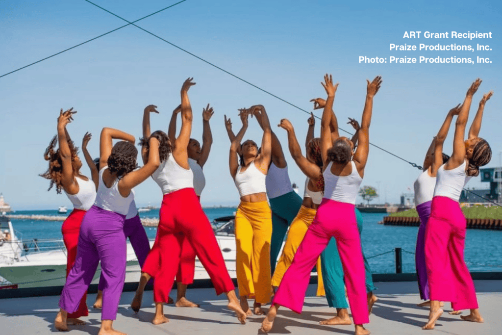 Ensemble of dancers from ART grant recipient Praize Productions, Inc. in synchronized performance with arms raised to the sky, wearing white crop tops and vibrant wide-leg pants in purple, red, yellow, pink, and teal, performing on waterfront stage with Lake Michigan visible in background