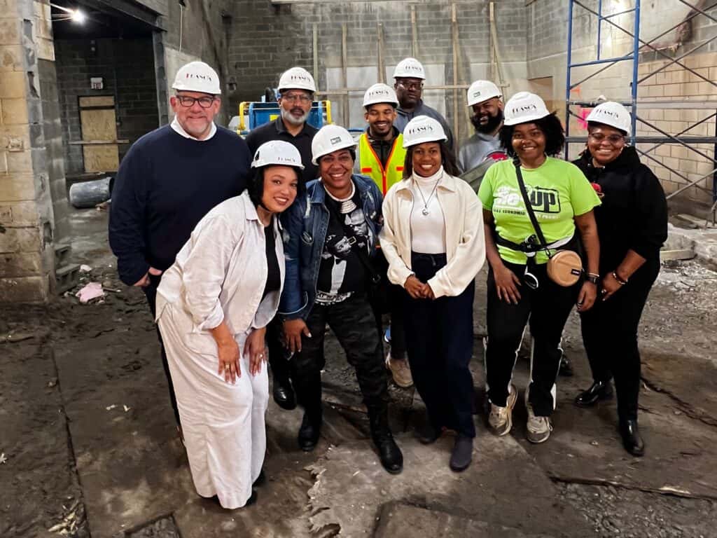 Ten members of the Field Foundation and R.A.G.E. teams wearing white hard hats pose together at the construction site of The Re-Up, 59th and Racine, amid exposed brick and scaffolding.