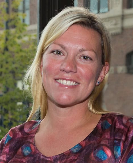 Headshot of Nora Daley smiling outdoors, wearing a purple and teal patterned top.