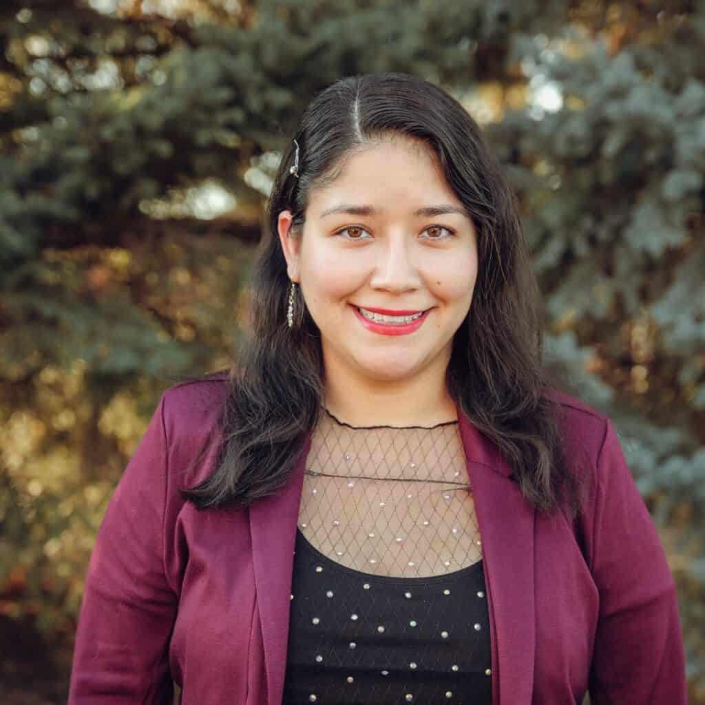 Adriana Velazquez, a woman with long dark hair, smiling in an outdoor headshot wearing a maroon blazer over a black top with sheer, jeweled detailing.
