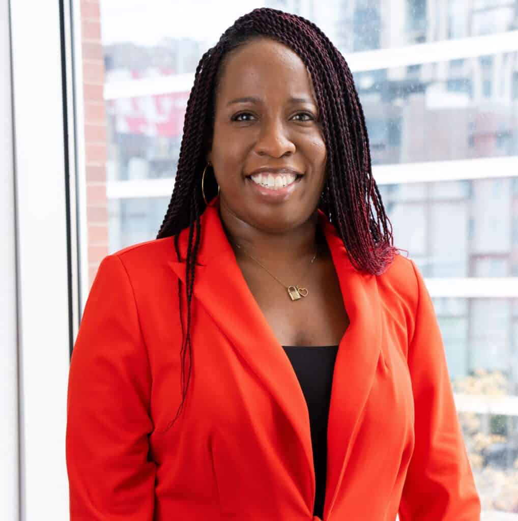 Candace Coleman, smiling in an orange blazer, with a city skyline visible through windows behind her.