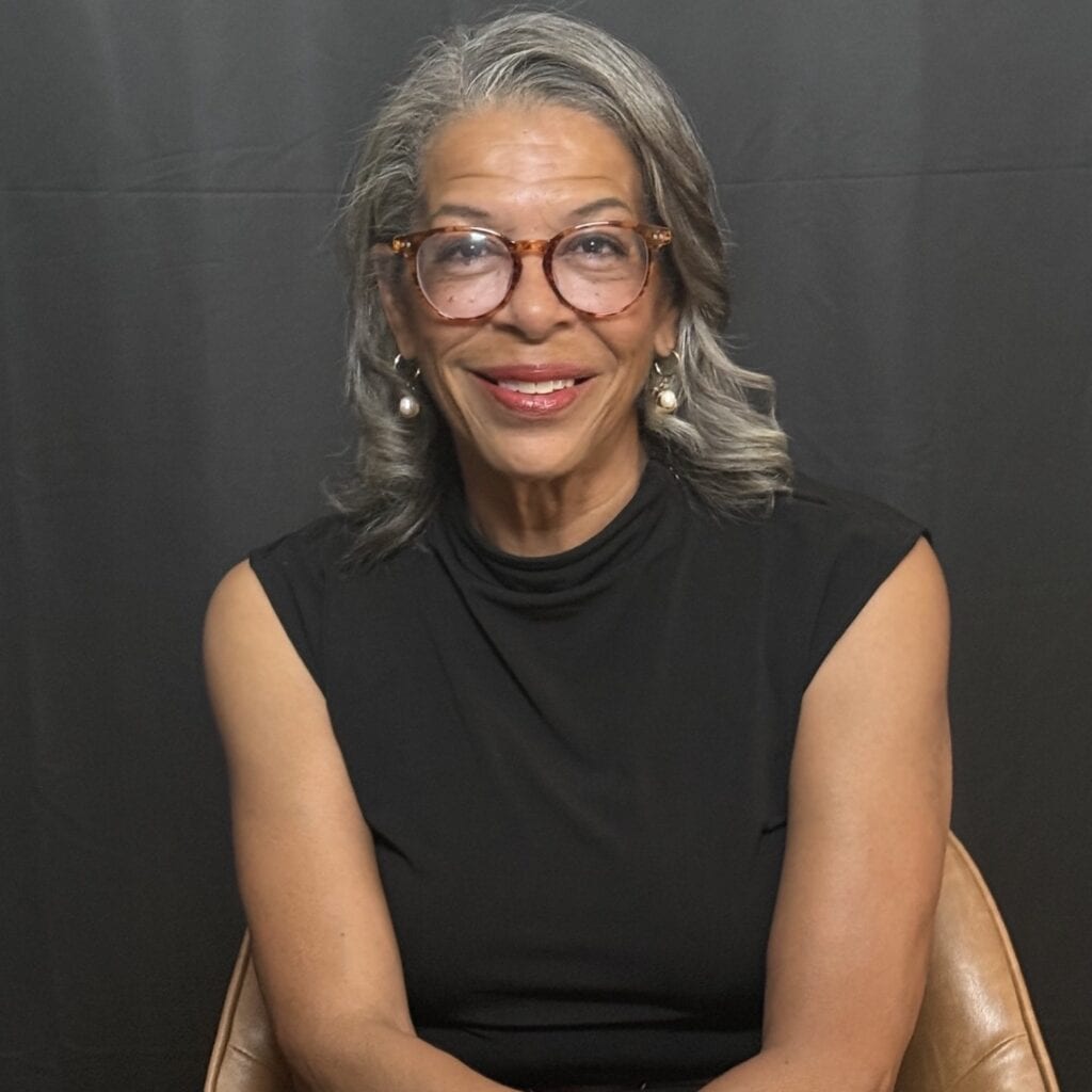 Cheryl Mainor Norman, seated and smiling in a black top, with glasses and gray hair, against a dark backdrop.