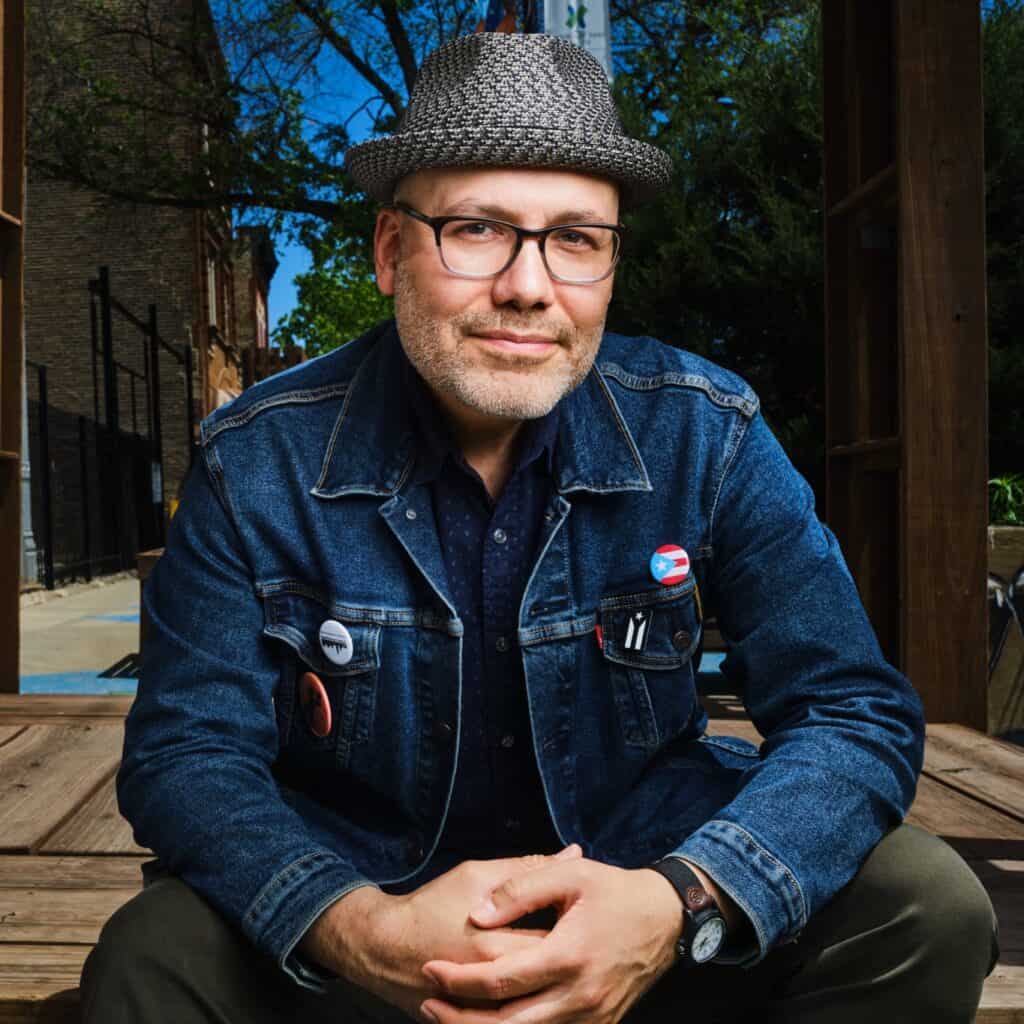 Ivan Vega, seated outdoors with hands clasped, wearing a denim jacket with pins including a Puerto Rican flag, a tweed hat, and glasses, smiling softly.