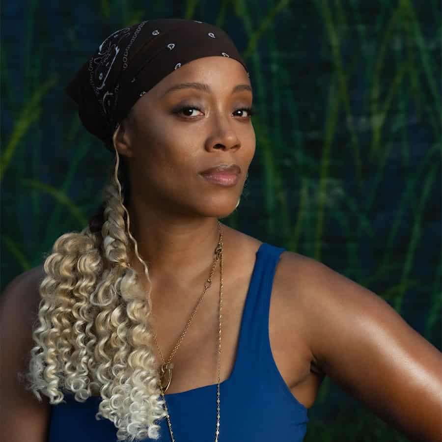 Janell Nelson posing confidently in a blue top with a long feather pendant necklace and a patterned headscarf, set against a dark green leaf-patterned backdrop.