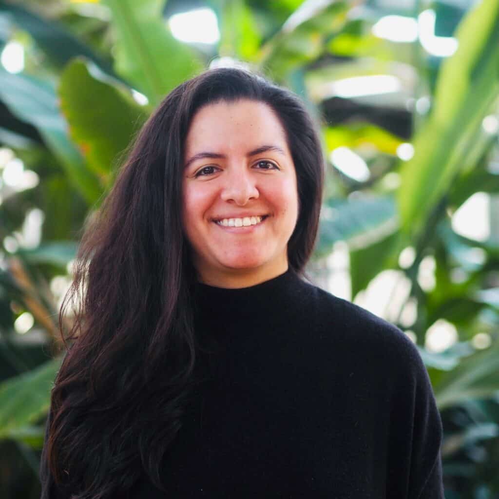 Xanat Sobrevila smiling in a black turtleneck, with lush green foliage in the background.