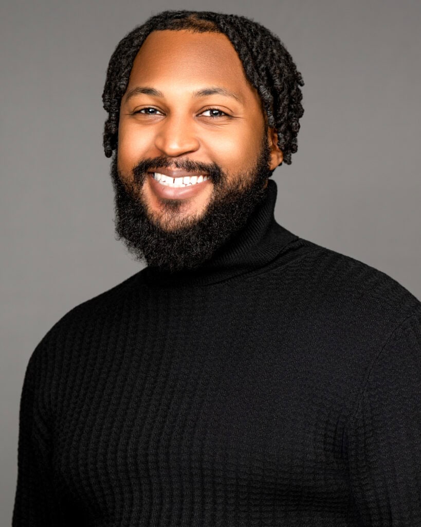 Headshot of Field Fellow Darius Caffey smiling, wearing a black turtleneck sweater against a gray background.